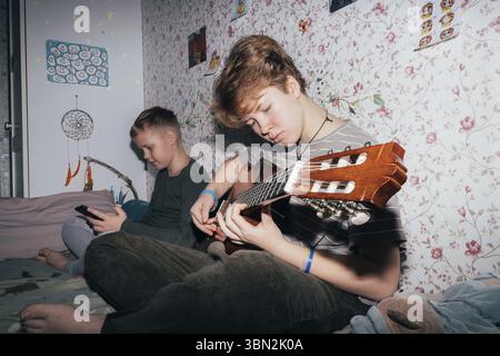 I bambini passano del tempo, la ragazza suona la chitarra, il ragazzo usa un telefono cellulare Foto Stock