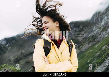 Una giovane donna rilassata torce la testa e i capelli lunghi soffiano al vento. Primo piano, ritratto di una donna in un impermeabile giallo in montagna Foto Stock