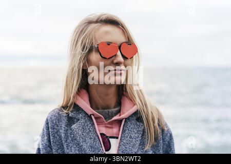 Primo piano di un viso femminile piacevole con cuori rossi di carta negli occhi invece di occhiali. Concetto di donna innamorata e festa di San Valentino Foto Stock