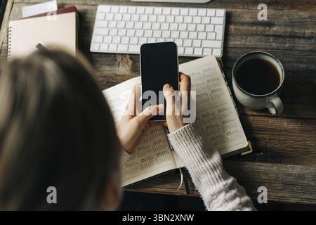 Draufsicht auf einen Schreibtisch, auf dem eine nicht erkennbare Frau ein Mobiltelefon und einen Organizer fuer die Arbeit benutzt Foto Stock