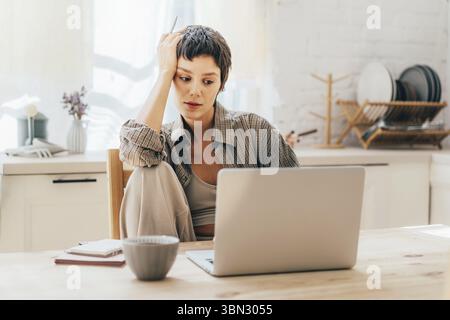 La giovane bruna donna seduta in cucina sul posto di lavoro utilizza un computer portatile per la comunicazione Foto Stock