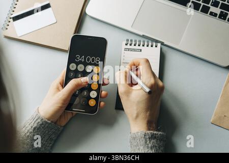 Draufsicht auf eine nicht erkennbare Frau, die auf einem Buerotisch rechnet und Notizen schreibt Foto Stock