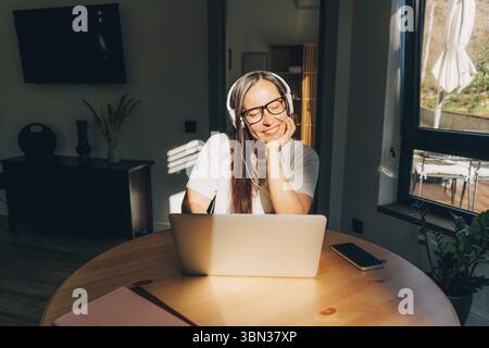 Junge, Glueckliche Frau, Die zu Hause am Tisch sitzt und im Sonnenlicht mit Kopfhoerern und laptop Musik hoert Foto Stock