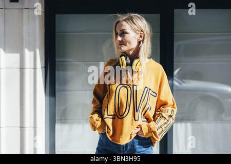 Ritratto urbano di giovane donna elegante in felpa casual gialla con gadget per l'ascolto di musica. Gli ampi auricolari wireless gialli sono appesi al collo Foto Stock