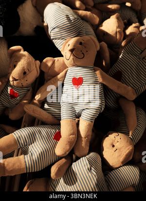 Simpatici orsetti di peluche che indossano abiti a righe blu con cuori rossi, disposti ordinatamente in un cestino dei giocattoli sotto una luce calda. Una vetrina di un peluche per bambini st Foto Stock