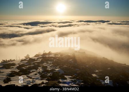 Alba sulla cima innevata della montagna. La calda luce dorata del sole che sorge bagna le nuvole in delicate sfumature di arancione e rosa. Neve e rocce sparse in primo piano aggiungono texture e profondità alla scena. Foto Stock