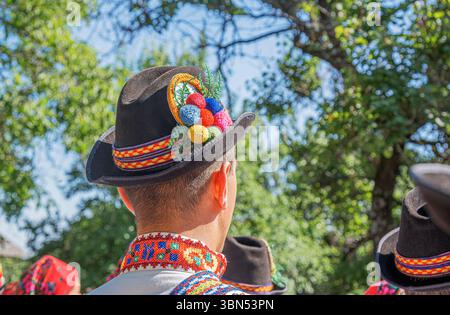 Un uomo in tradizionale abbigliamento folk, adornato con un cappello decorato con cura, si affaccia su una vivace scena all'aperto. Foto Stock