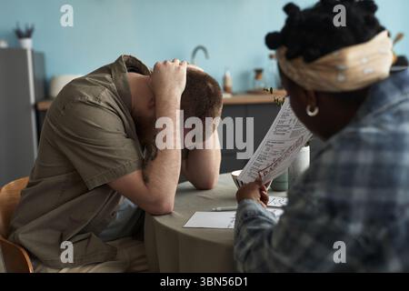 Giovane uomo adulto caucasico seduto al tavolo tenendo la testa tra le mani mentre la giovane donna adulta di colore seduta di fronte a un documento, entrambi sembrano impegnati in un'intensa discussione Foto Stock