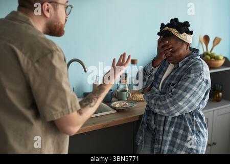 Giovane uomo adulto caucasico che gestiva con rabbia verso la giovane donna adulta nera in piedi accanto al bancone della cucina che copre il viso con la mano, coppia che vive conflitti o discussioni a casa Foto Stock