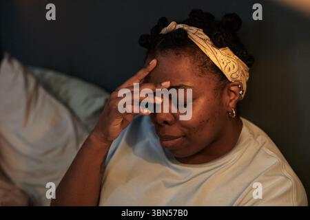 Donna di colore giovane adulto seduto sul letto toccando la fronte a occhi chiusi che mostrano stress o frustrazione durante situazioni di conflitto, testa leggermente inclinata, con archetto Foto Stock