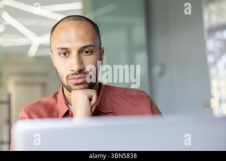 Un uomo pensivo, dalla pelle scura e con la barba appoggia il mento sulla mano davanti a un portatile, perso nel pensiero. Foto Stock