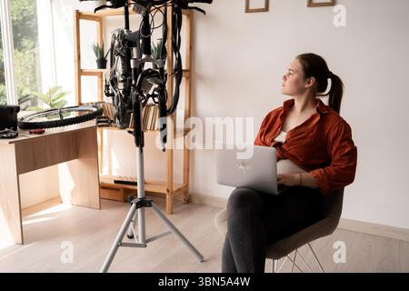 Giovane donna in abiti casual con PC portatile che ripara la bicicletta a casa. Copia spazio Foto Stock