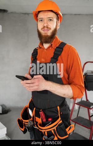 Giovane costruttore maschile barbuto con smartphone e utensili da lavoro in cintura in piedi in una nuova casa. Tecnologia, riparazione, costruzione e concetto di costruzione. Foto Stock