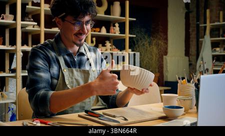 Uomo caucasico studente classe online laboratorio di ceramica blogger maschio artigiano artista designer potter insegnante che mostra ciotola fatta a mano modellando argilla Foto Stock