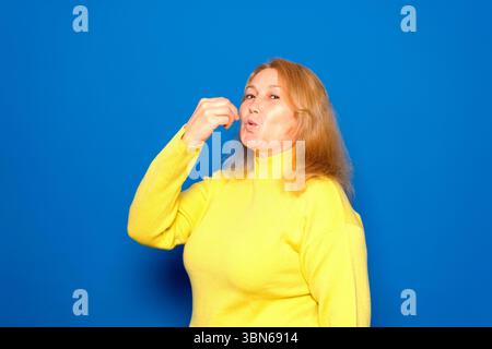 Donna caucasica di 40 anni che indossa un maglione giallo con un sorriso positivo e felice che mostra un delizioso segno gourmet perfetto isolato sul blu Foto Stock