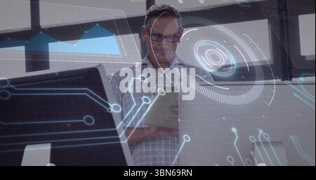 Revisione di un uomo che indossa una camicia a scacchi che esamina i dati del tablet alla scrivania dell'ufficio con monitor e ologrammi Foto Stock