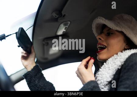 Una donna elegante applica il rossetto rosso in un'auto parcheggiata utilizzando lo specchietto retrovisore. Indossa un comodo cappello e un cappotto invernale, catturando ogni momento Foto Stock