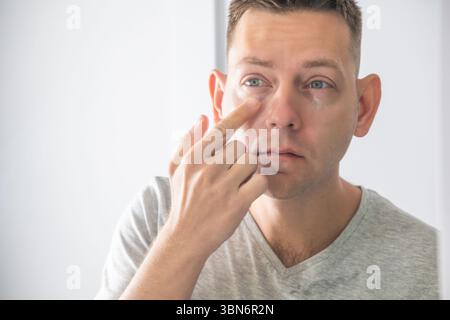 Uomo che applica crema per gli occhi sotto gli occhi davanti allo specchio per la cura della pelle. Concetto di bellezza e cura della pelle. Cura del viso maschile, trattamenti viso, anti Foto Stock