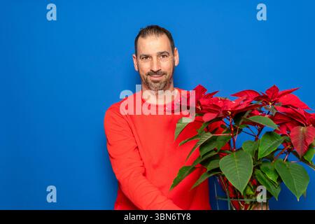 Uomo ispanico barbuto di 40 anni che indossa un maglione rosso con una graziosa pianta di Natale spagnola. Isolato su sfondo blu. Foto Stock