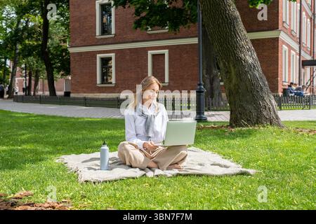 Donna impegnata con laptop per film, videochiamata all'aperto sul prato del parco cittadino con una bottiglia d'acqua Foto Stock