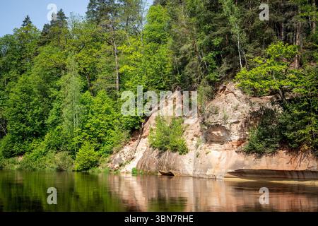 Scogliera rocciosa di arenaria che si riflette nell'acqua del fiume circondata da una fitta foresta verde in estate. Foto Stock