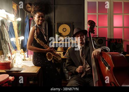 Donna nera giovane adulta che tiene il sassofono seduto accanto a un uomo caucasico di mezza età che suona il contrabbasso nel camerino del jazz club, entrambi i musicisti si preparano per l'esibizione Foto Stock