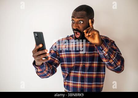 Uomo nero sorpreso e scioccato con una camicia accogliente davanti al muro e videochiamata sullo smartphone. Foto Stock