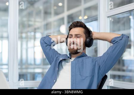 Un uomo rilassato che indossa le cuffie sorride mentre è seduto in ufficio con le mani dietro la testa. Foto Stock