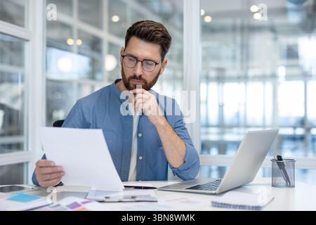 Un uomo con una camicia blu e un paio di occhiali esamina i documenti, considerando attentamente le informazioni. Foto Stock