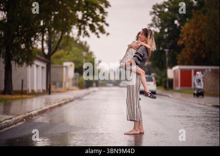 Donna che tiene il bambino in braccio su strada bagnata durante la pioggia, a piedi nudi e gioiosa in un tenero momento di connessione. Foto Stock