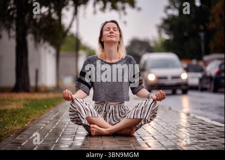 La donna siede a gambe incrociate, gli occhi chiusi, meditando pacificamente sotto la pioggia sul marciapiede della città, abbracciando calma e consapevolezza. Foto Stock