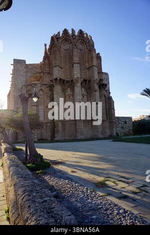 Cipro, Famagosta, Cattedrale di San Nicola, Moschea Lala Mustafa Pasha, chiesa medievale convertita in moschea dai turchi ottomani nel 1571, con un minareto che si affaccia sul mare. Grande chiesa in pietra ornata con uno stile architettonico gotico, che si erge maestosamente su un cielo azzurro. La facciata della chiesa è adornata da intricate incisioni e presenta numerose finestre ad arco e porte. La prospettiva dell'immagine è da un'angolazione bassa, sottolineando l'imponente presenza della chiesa. La chiesa si trova in un cortile lastricato di pietra di colore chiaro, e un muro di pietra con un albero e un lampione Foto Stock