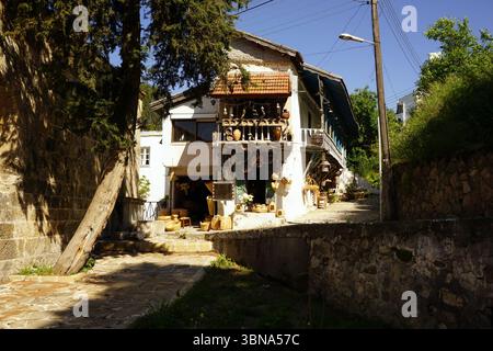 Cipro, Edremit, Una pittoresca scena cittadina, con un edificio bianco a due piani caratterizzato da un tetto in piastrelle rosse e un balcone adornato da una ringhiera di legno. L'edificio si trova su un sentiero di pietra, con un muro di pietra sul lato destro. Un albero si trova sul lato sinistro del percorso, gettando un'ombra sul terreno. L'edificio ha anche un piccolo negozio o negozio al piano terra. Il cielo è blu chiaro e le linee elettriche corrono lungo la parte superiore dell'immagine. La scena e' inondata dalla luce del sole, creando un'atmosfera calda e invitante. L'occhio di un artista e la sua immaginazione Foto Stock