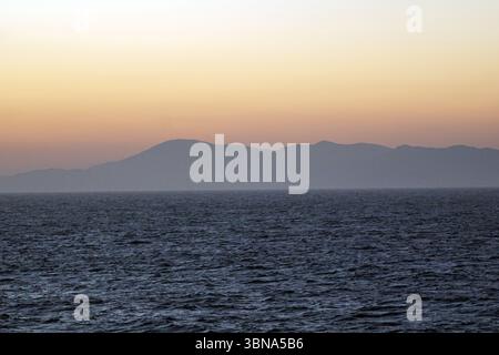 Isole greche tra Cipro e Atene, Un tranquillo paesaggio costiero al tramonto. Il cielo è dipinto in tonalità arancio e rosa, con il sole che tramonta dietro una catena di montagne in lontananza. Le montagne sono avvolte da una nebbia nebulosa, creando un senso di profondità e mistero. L'oceano, di colore grigio-blu scuro, occupa la parte inferiore dell'immagine, con piccole onde che si infrangono dolcemente contro la costa. La linea dell'orizzonte è chiaramente visibile, separando l'oceano dal cielo. Le montagne sembrano essere una catena di colline o basse montagne, piuttosto che una serie di cime distinte. L'occhio di un artista e.. Foto Stock