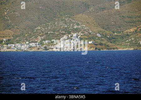 Isole greche tra Cipro e Atene, Una pittoresca cittadina annidata su una collina, con edifici bianchi sparsi nel paesaggio. Il paese è situato su una collina che degrada verso il mare blu profondo, creando un netto contrasto tra le strutture artificiali e l'ambiente naturale. La collina è ricoperta di vegetazione, tra cui alberi e arbusti, e sembra essere un mix di vegetazione e forse alcune aree coltivate. Il mare è di colore blu intenso, con piccole onde che si infrangono dolcemente sulla riva. La prospettiva dell'immagine è dal mare, guardando verso la città, Foto Stock