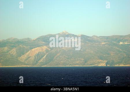 Isole greche tra Cipro e Atene, Un paesaggio montuoso con una cima prominente al centro, caratterizzato da una piccola struttura o antenna sulla sua sommità. Le montagne sono principalmente di colore verde e marrone tenue, con alcune aree di tonalità più chiare, abbronzate o beige. Il cielo è azzurro e limpido. La prospettiva è dal mare, con l'oceano blu scuro che si estende in primo piano. Le montagne sembrano essere relativamente pianeggianti o leggermente inclinate nelle aree in cui sono visibili. L'occhio di un artista e la sua immaginazione Foto Stock