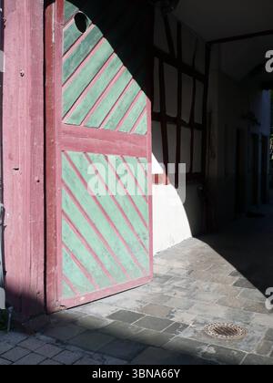 Idstein, Germania, Una porta in legno con una vivace cornice rossa e un suggestivo motivo a strisce verdi e bianche. La portiera è leggermente socchiusa, a rivelare un'occhiata all'interno. La porta è adagiata su una parete bianca con una sezione di travi di legno esposte e una parte di una struttura è visibile a destra. Il terreno di fronte alla porta è pavimentato con pietre grigie e un tombino è visibile in primo piano. La scena è immersa nella luce del sole, proiettando ombre sulla porta e sul muro, creando un contrasto tra aree chiare e scure. L'occhio di un artista e la sua immaginazione Foto Stock