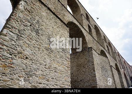 Kavala, Grecia, Un acquedotto in pietra costruito con grandi pietre di forma irregolare nelle sfumature di grigio e marrone. L'acquedotto presenta archi multipli e una serie di archi collegati, creando una struttura continua. La prospettiva è da un angolo basso, guardando verso l'alto l'altezza torreggiante dell'acquedotto. Un uccello viene catturato a metà volo contro un cielo azzurro con alcune nuvole bianche. L'immagine è priva di testo o oggetti creati dall'uomo, concentrandosi esclusivamente sulla struttura in pietra naturale e sull'uccello in volo. L'occhio di un artista e la sua immaginazione Foto Stock