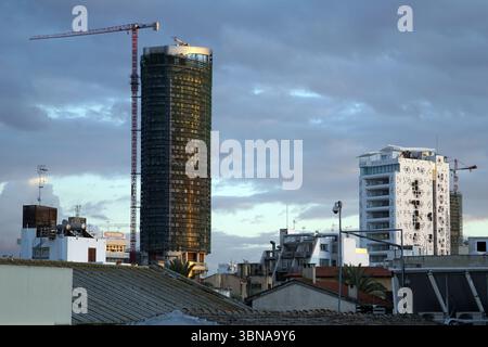Nicosia (Lefkosia), Cipro, l'ultima capitale divisa del mondo, Un paesaggio urbano al crepuscolo, con un alto edificio cilindrico in costruzione, la sua facciata scura illuminata dal sole che tramonta. L'edificio è circondato da altre strutture, tra cui un edificio bianco con un tetto blu e un edificio bianco con un tetto rosso. Una gru rossa è visibile sul lato sinistro dell'immagine e un edificio bianco con un tetto blu è sulla destra. Il cielo è pieno di nuvole, che proiettano una luce soffusa e diffusa sulla scena. L'immagine viene scattata da un alto punto panoramico, fornendo una vista completa dello skyline. An Foto Stock
