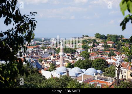 Plovdiv, la seconda città più grande della Bulgaria, è una delle città più antiche d’Europa continuamente abitate, con una storia che abbraccia oltre 8.000 anni. Incastonato lungo il fiume Maritsa, Un paesaggio urbano con una moschea con un alto minareto e diverse cupole, situato al centro della cornice. Il paesaggio urbano è un mix di edifici con tetti rossi e arancioni, intervallati da alberi e vegetazione. La città si estende in lontananza, con una collina o un'area sopraelevata visibile sullo sfondo. Il cielo è azzurro con alcune nuvole sparse. L'immagine viene scattata da un alto punto panoramico che offre una vista panoramica Foto Stock