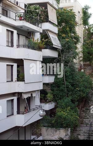 Atene, la vibrante capitale della Grecia, immersa nella storia e nella cultura, è la culla della civiltà e della democrazia occidentali. Un edificio di appartamenti bianchi a più piani con balconi su ogni piano. L'edificio presenta un mix di balconi con ringhiere nere e tende, alcuni dei quali sono chiusi. I balconi sono adornati con piante e fiori in vaso, aggiungendo un tocco di verde al paesaggio urbano. Una porzione di un secondo edificio è visibile dietro la struttura principale. L'edificio e' circondato da vegetazione lussureggiante, che include alberi e cespugli, e una serie di scale conduce all'edificio. A. Foto Stock