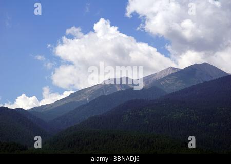 Una maestosa catena montuosa sotto un cielo blu limpido punteggiato da soffici nuvole bianche. Le montagne, con le loro aspre vette e valli, sono coperte da un denso strato di alberi verdi scuri. Le montagne sembrano essere un mix di roccia e vegetazione, con alcune aree che mostrano segni di erosione o di agenti atmosferici. La prospettiva dell'immagine è da un'angolazione bassa, guardando verso l'alto verso le montagne torreggianti, creando un senso di soggezione e grandezza. Il cielo è un blu vibrante, che contrasta splendidamente con il verde scuro delle montagne e il bianco delle nuvole. Didascalia a forma di occhio e immaginazione di un artista, Bansko i. Foto Stock