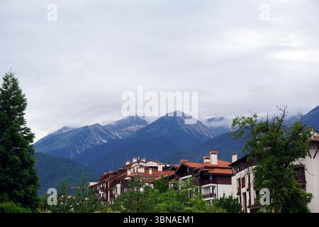 Un tranquillo paesaggio montano con un gruppo di edifici annidati in primo piano. Gli edifici, con i loro tetti rossi e le pareti bianche, sono situati su una leggera pendenza, affacciati sulle maestose montagne. Le montagne, ricoperte da lussureggianti alberi verdi, si innalzano maestosamente sullo sfondo, le loro vette parzialmente oscurate da uno strato di nuvole. Il cielo è grigio chiaro, e suggerisce una giornata ricoperta. L'immagine viene scattata da una prospettiva leggermente rialzata, fornendo una visione completa della scena. Didascalia a forma di occhio e immaginazione di un artista. Bulgaria, Bansko, Pirin Mountains Foto Stock