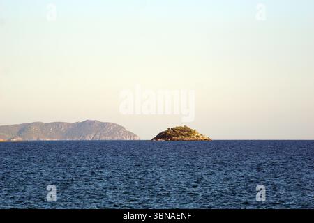 Una tranquilla scena di una piccola isola circondata da un vasto e profondo oceano blu, vicino a Bozyazi, Turchia. L'isola è coperta da una lussureggiante vegetazione verde e ha una costa rocciosa. In lontananza, una grande montagna rocciosa sorge dall'orizzonte, con la sua superficie aspra in contrasto con il mare calmo. Il cielo sopra è un azzurro chiaro e pallido, privo di nuvole. L'immagine viene scattata da lontano, fornendo una vista completa dell'isola e dell'ambiente circostante. L'occhio di un artista e la sua immaginazione Foto Stock