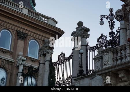 Budapest, Ungheria: L'incantevole Perla del Danubio, la collina del castello di Budapest. Una parte di un edificio dallo stile architettonico classico, caratterizzato da una combinazione di elementi in pietra e metallo. La facciata dell'edificio è principalmente in pietra, con dettagli ornati e un balcone al piano superiore. Il balcone ha una balaustra e un elemento decorativo. L'edificio presenta anche un cancello in ferro battuto nero con elementi decorativi e un insieme di colonne con capitelli ornati. Un lampione stradale con base decorativa è visibile sul lato destro dell'immagine. Il cielo è azzurro e l'immagine lo è Foto Stock
