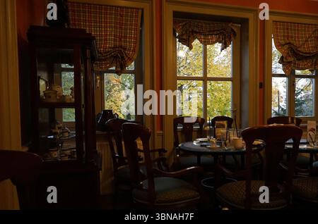 Budapest, Ungheria: L'incantevole Perla del Danubio, Una sala da pranzo accogliente con un'atmosfera calda e invitante. La camera presenta un tavolo in legno circondato da sei sedie in legno abbinate con tappezzeria marrone scuro o nera e cuscini a motivi geometrici. Il tavolo e' allestito con piatti, bicchieri e argenteria, suggerendo che un pasto sta per essere servito. La sala è illuminata da luce naturale che scorre attraverso tre finestre con cornici bianche e tende color crema. Le tende sono adornate con un motivo a quadri in sfumature di rosso, verde e crema. Le pareti sono dipinte di un caldo colore arancione e scuro Foto Stock