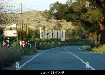Viaggiando a Cipro, Una strada tortuosa che curva dolcemente a destra, circondata da un lussureggiante paesaggio di alberi e arbusti. La strada è segnata da linee bianche e presenta alcune strisce rosse e bianche lungo il bordo della strada. Un cartello sul lato sinistro della strada indica la direzione per "Benvenuti". La strada è delimitata da una bassa area erbosa sulla sinistra e da una collina con un mix di vegetazione e alberi sulla destra. Il cielo è azzurro e il sole splende brillantemente, proiettando un caldo bagliore sulla scena. L'occhio di un artista e la sua immaginazione Foto Stock