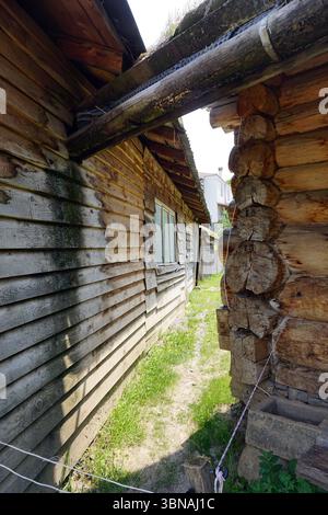 Una struttura rustica in legno, possibilmente una capanna in legno, con un aspetto invecchiato e intempestivo. L'edificio presenta un tetto inclinato e uno sbalzo in legno sostenuto da una trave in legno. Una piccola finestra è visibile sul lato dell'edificio. Una recinzione o ringhiera di legno corre lungo il lato dell'edificio. Di fronte all'edificio è visibile un sentiero sterrato o una passerella che conduce verso un'area erbosa. Una porzione di un secondo edificio è visibile sullo sfondo. La scena è ambientata contro un cielo azzurro, con una piccola sezione di un edificio bianco visibile in lontananza. Didascalia a forma di occhio e immaginazione di un artista. Foto Stock