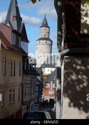 Idstein, Germania, Una vista pittoresca, con un'importante torre del castello che sorge sullo sfondo. La torre, costruita in pietra di colore chiaro, presenta un tetto grigio scuro e una piccola bandiera sulla sommità. La scena è annidata tra edifici con un mix di stili architettonici, tra cui case in legno con tetti marrone scuro o neri e pareti color crema. Gli edifici sono principalmente alti due o tre piani, con alcuni che hanno finestre multiple. Alcune persone possono essere viste camminare per la strada di fronte agli edifici. Il cielo è blu chiaro con alcune nuvole sparse. Una porzione di colore scuro Foto Stock