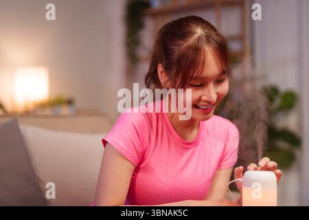 Una donna asiatica adulta sorride mentre utilizza un diffusore di oli essenziali aromaterapici a casa sua per godere di benessere e longevità e creare un relax Foto Stock
